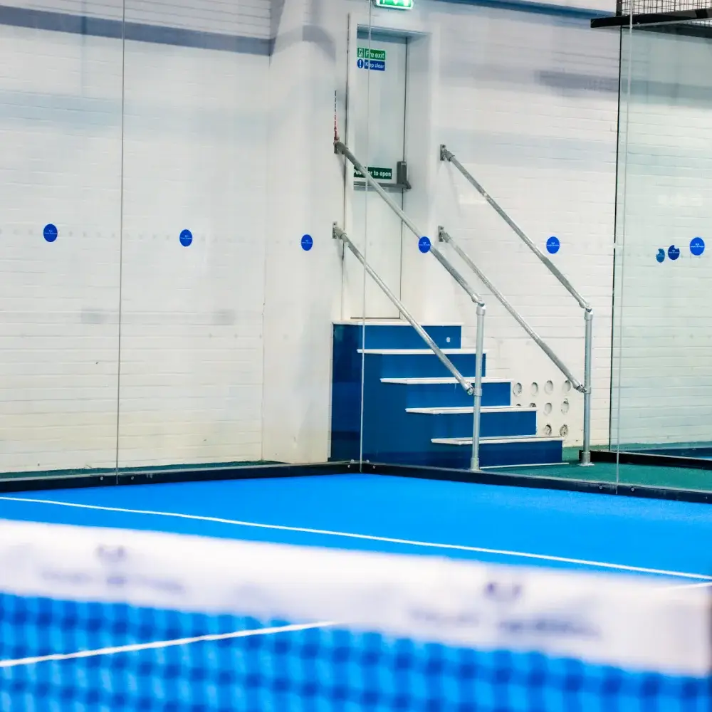 Indoor padel court corner with blue surface and glass wall at Court De Padel